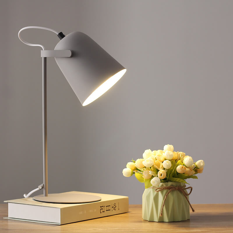 Graue Tischlampe leuchtet auf einem Buch vor grauer Wand, daneben eine Vase mit hellen Röschen auf einem Holztisch. 10