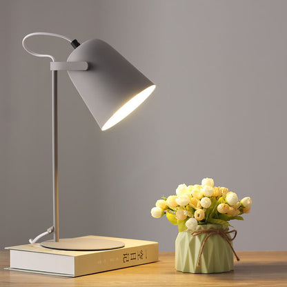 Graue Tischlampe leuchtet auf einem Buch vor grauer Wand, daneben eine Vase mit hellen Röschen auf einem Holztisch. 10