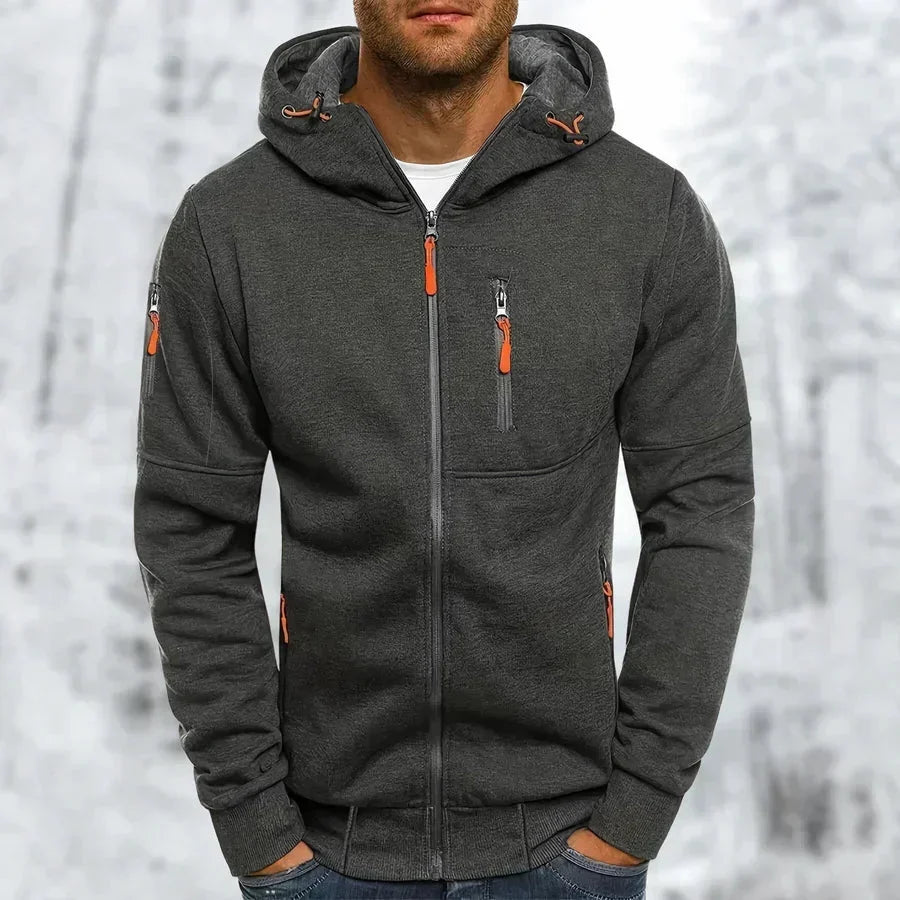 Mann trägt den dunkelgrauen Kapuzenpullover mit orangefarbenen Zippern, aufgesetzter Brusttasche und Rippbündchen vor verschneitem Wald. 9