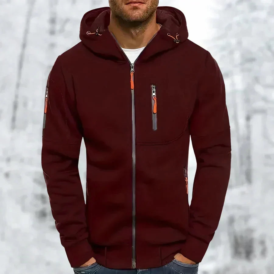 Mann im weinroten Kapuzenpullover mit orangefarbenen Reißverschlusszügen und grauem Zipper, vor verschneiter Waldkulisse. 4