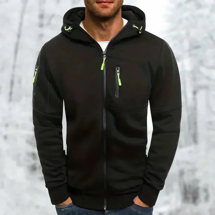 Mann trägt den schwarzen Kapuzenpullover mit neongrünen Zippern und Ärmeltasche, Hände in den Hosentaschen, vor Winterkulisse. 7