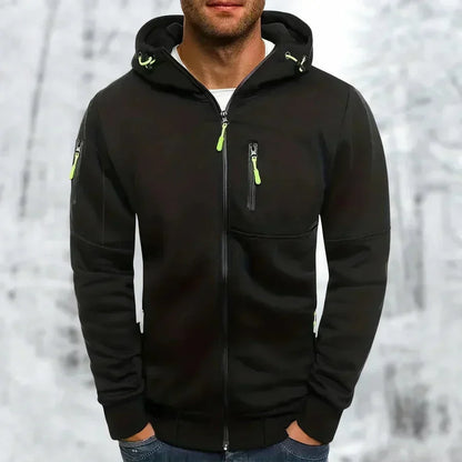 Mann trägt den schwarzen Kapuzenpullover mit neongrünen Zippern und Ärmeltasche, Hände in den Hosentaschen, vor Winterkulisse. 7