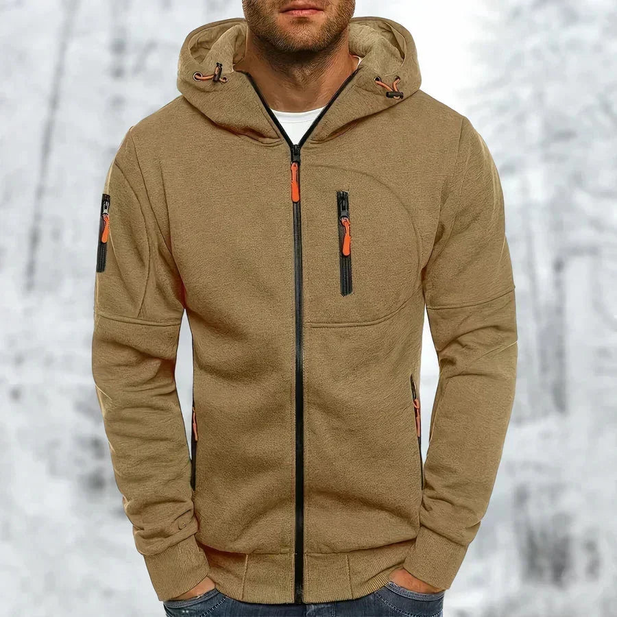 Mann im khakibraunen Kapuzenpullover mit schwarzem Reißverschluss, orangefarbenen Zugbändern und Ärmeltasche vor winterlichem Hintergrund. 10