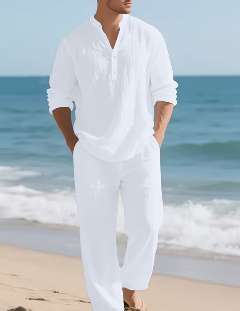 Mann in weißem Henley-Hemd mit Knopfleiste und weiter weißer Hose, Hände in den Taschen, am Sandstrand vor dem Meer. 2