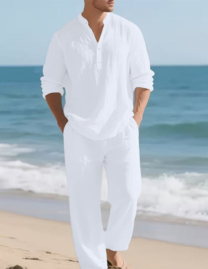 Mann in weißem Henley-Hemd mit Knopfleiste und weiter weißer Hose, Hände in den Taschen, am Sandstrand vor dem Meer. 2