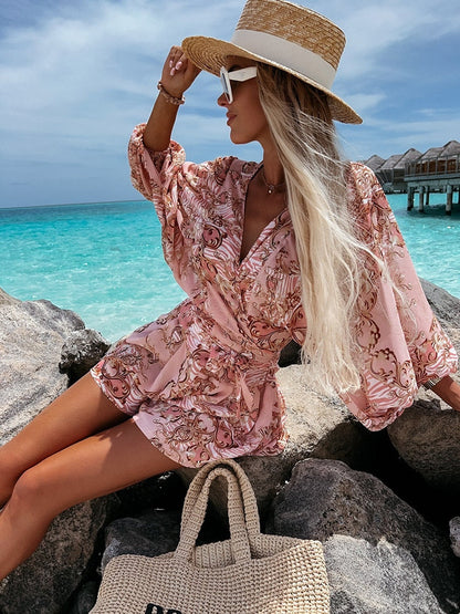 Frau in rosa Sommerkleid und Strohhut am Strand, sitzt auf Felsen, türkisfarbenes Meer im Hintergrund.