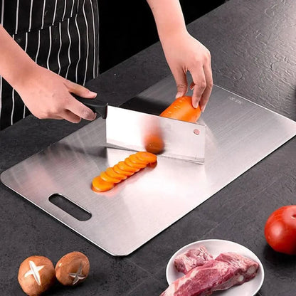 Person schneidet mit einem Hackbeil Möhrenscheiben auf dem gebürsteten Metallbrett, daneben Pilze, Fleisch auf einem Teller und eine Tomate. 3