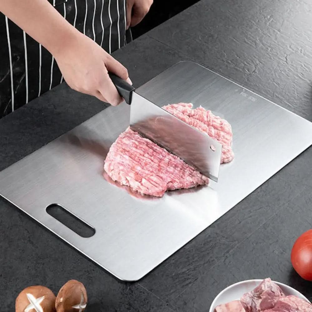 Metallisches Schneidebrett mit Grifföffnung auf dunkler Arbeitsplatte, auf dem eine Person mit Hackbeil rosa Hackfleisch zerkleinert. 1