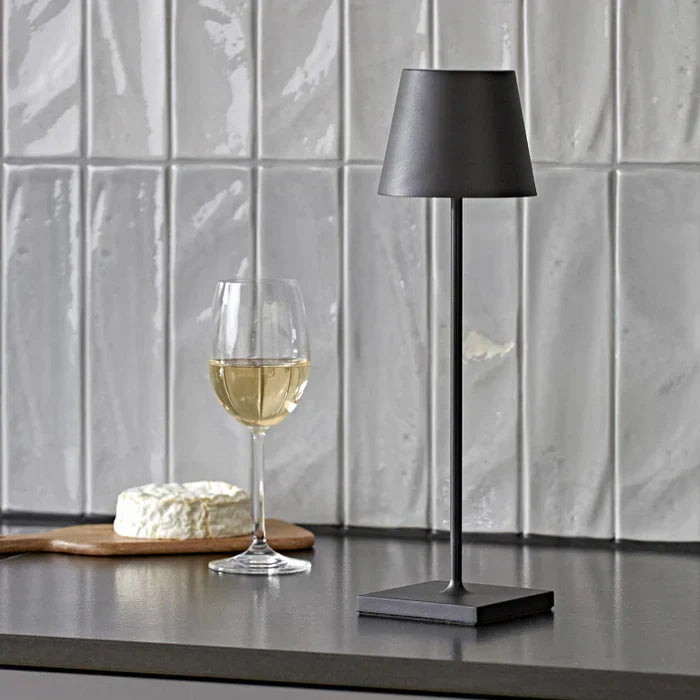 Schwarze Tischlampe aus Metall neben Weinglas und Käse auf grauem Küchentresen vor Fliesenwand.