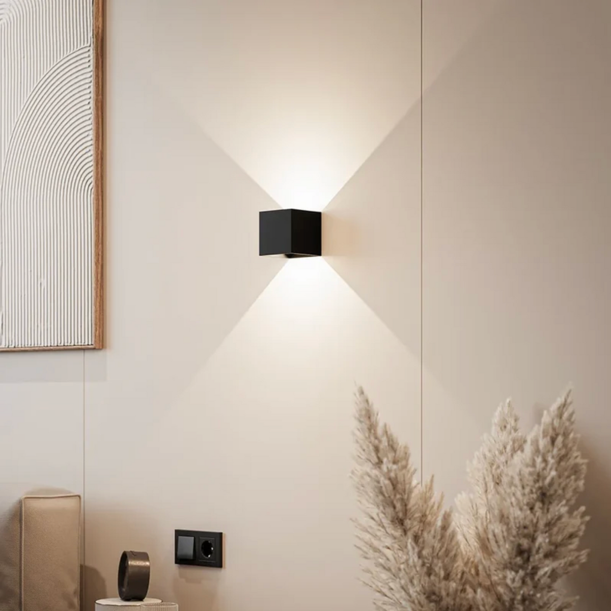 Schwarze würfelförmige Wandleuchte strahlt Licht nach oben und unten an einer beigen Wand neben Pampasgras und Wandbild. 1