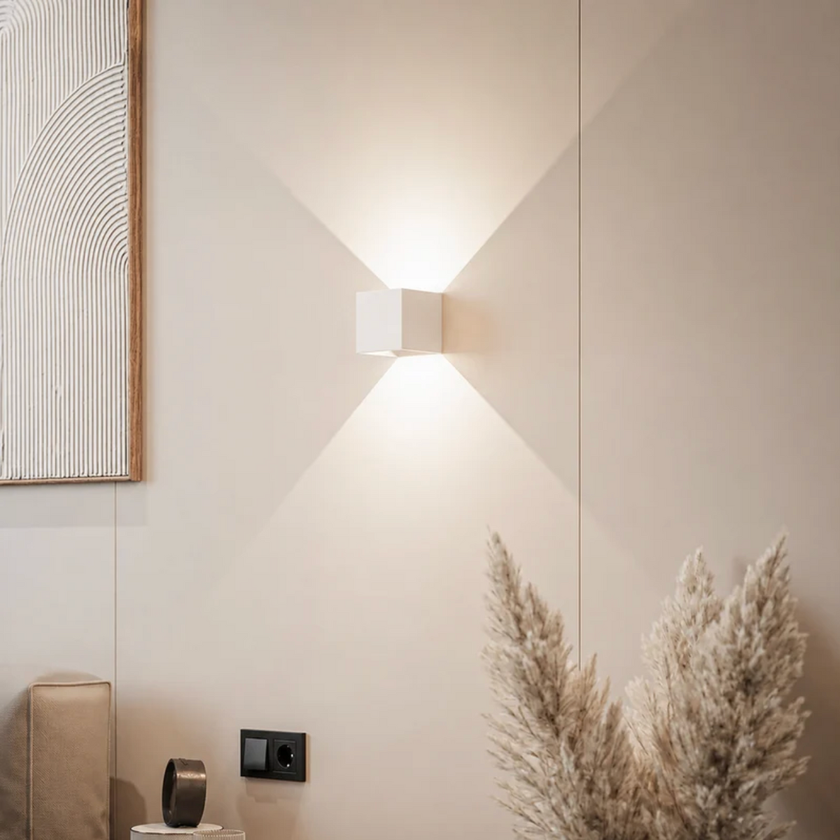 Weiße Würfelleuchte an beiger Wand mit Lichtkegeln nach oben und unten, daneben Pampasgras, Wandbild und Steckdose. 12
