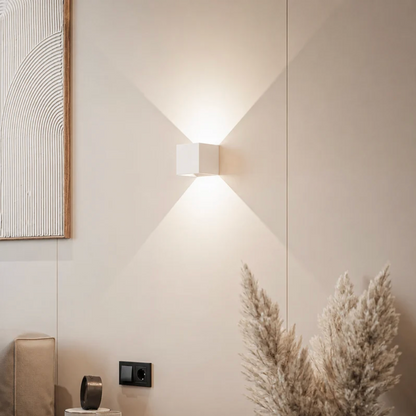 Weiße Würfelleuchte an beiger Wand mit Lichtkegeln nach oben und unten, daneben Pampasgras, Wandbild und Steckdose. 12