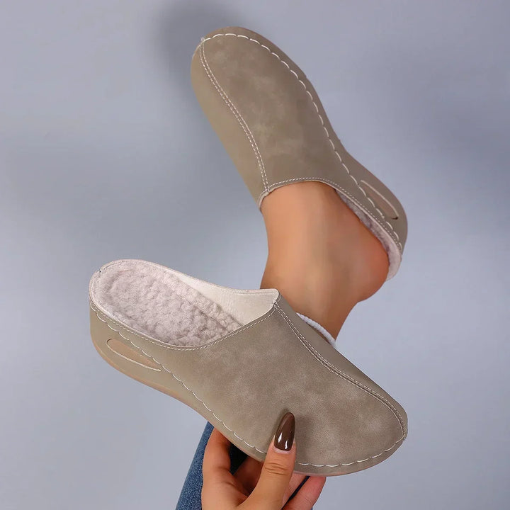 Beigefarbene Pantoffeln mit flauschigem Innenfutter, von einer Hand gehalten, daneben ein Fuß im zweiten Schuh. 6