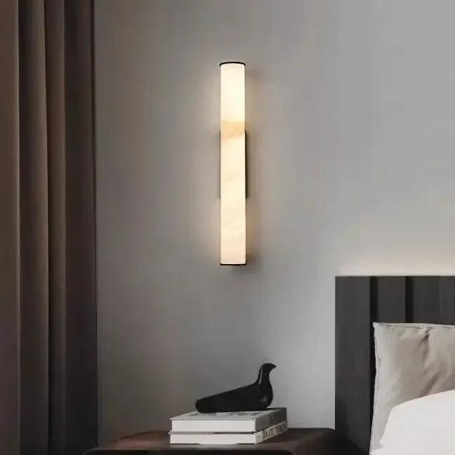 Schmale leuchtende Wandlampe an grauer Wand über einem Nachttisch mit schwarzer Vogelfigur und Büchern, neben Vorhang und Bett. 14