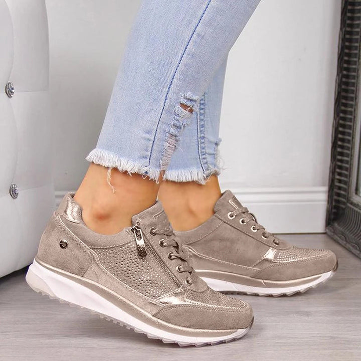 Sneaker in Taupe mit seitlichem Reißverschluss, Schnürung und glitzernden Details, getragen zu hellblauer ausgefranster Jeans vor weißer Polstermöbelwand. 1