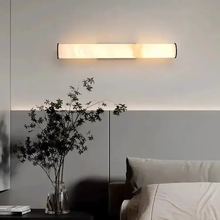Waagerecht montierte Wandlampe mit marmoriertem Zylinder über einem Bett, daneben ein Nachttisch mit Glasvase und dunklem Zweig. 16