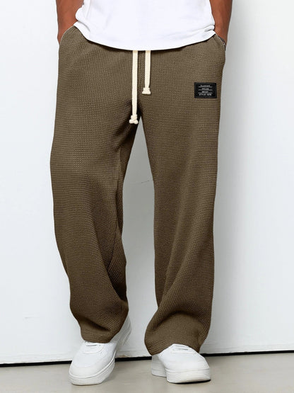 Waffelstrickhose in Braunoliv mit weitem Bein und beigem Kordelzug, Frontansicht am Model mit weißem Shirt. 5