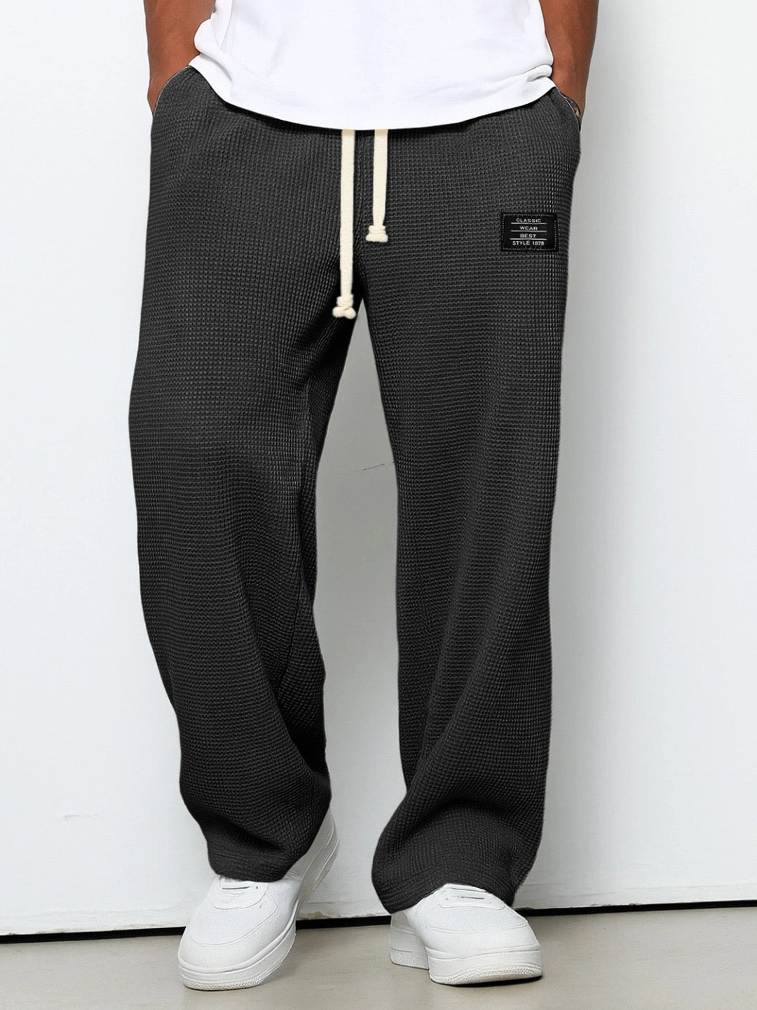 Weit geschnittene Waffelstrickhose in Dunkelgrau, getragen mit weißem T-Shirt und weißen Sneakern, Frontansicht mit hellem Kordelzug. 1