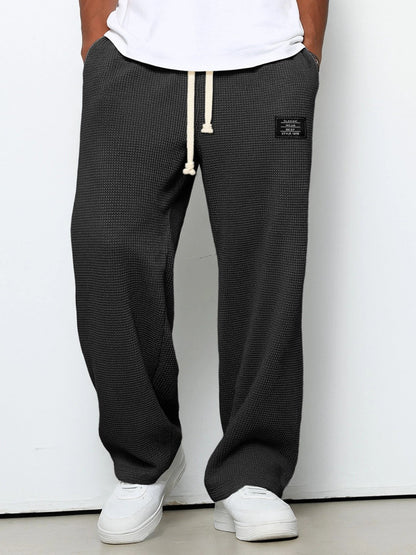 Weit geschnittene Waffelstrickhose in Dunkelgrau, getragen mit weißem T-Shirt und weißen Sneakern, Frontansicht mit hellem Kordelzug. 1
