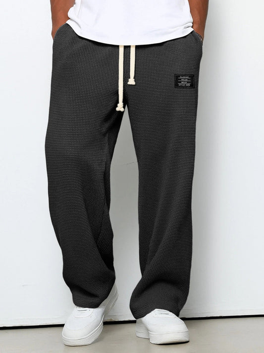 Weit geschnittene Waffelstrickhose in Dunkelgrau, getragen mit weißem T-Shirt und weißen Sneakern, Frontansicht mit hellem Kordelzug. 1