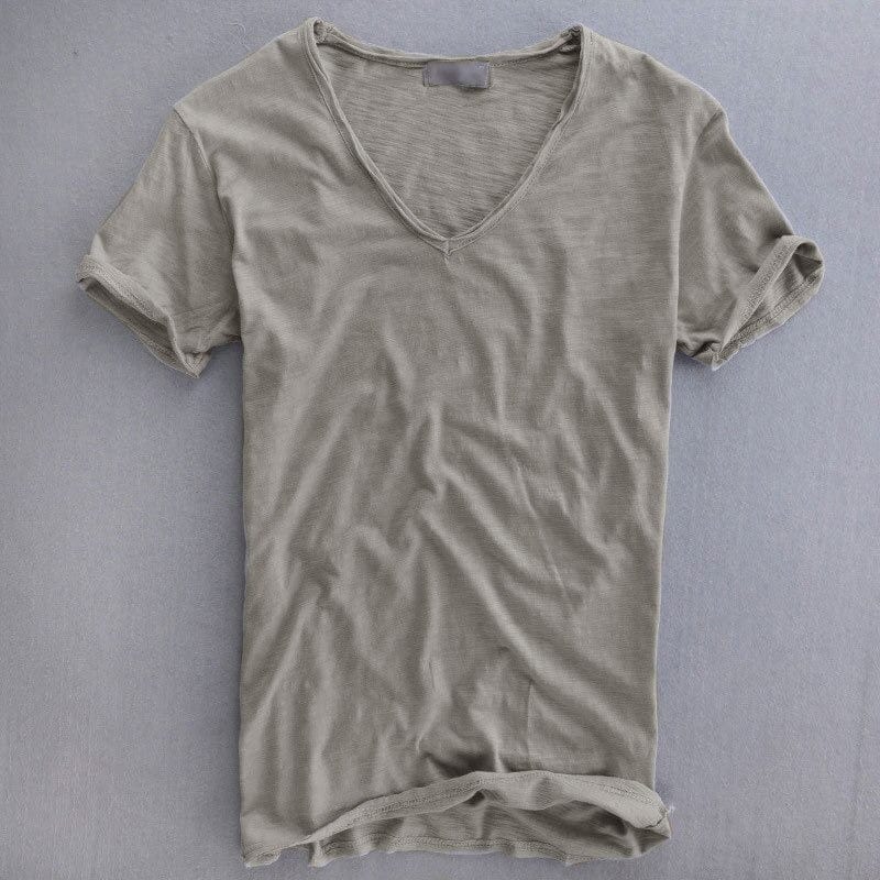 Kurzärmeliges T-Shirt mit tiefem V-Ausschnitt in Graubeige, flach auf grauem Untergrund ausgebreitet, Ärmel leicht umgeschlagen. 1