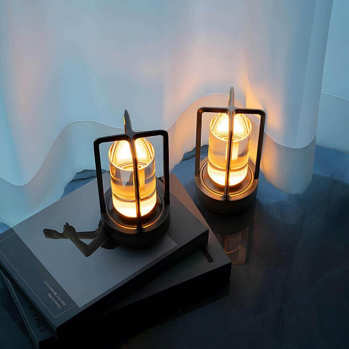 Zwei warm leuchtende Tischlampen mit dunklem Rahmen stehen auf einem Bücherstapel und dem Boden vor einem weißen Vorhang. 3