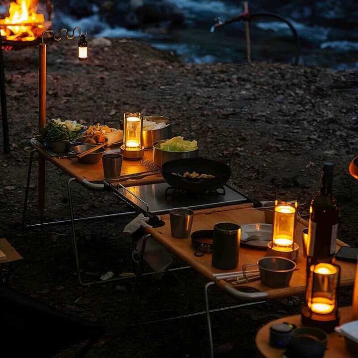 Mehrere Lampen beleuchten abends einen Campingtisch mit Kochgeschirr, Gaskocher und Weinflasche, im Hintergrund ein Lagerfeuer. 4
