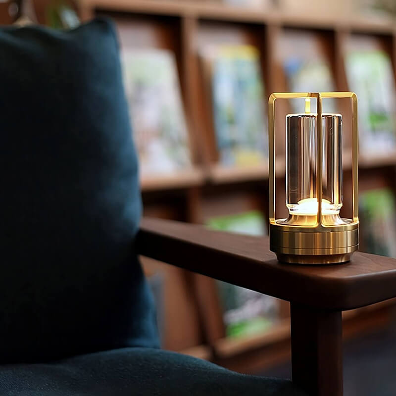 Messingfarbene Tischlampe leuchtet auf der Holz-Armlehne eines Sessels mit dunkelblauem Kissen vor einem Bücherregal. 8
