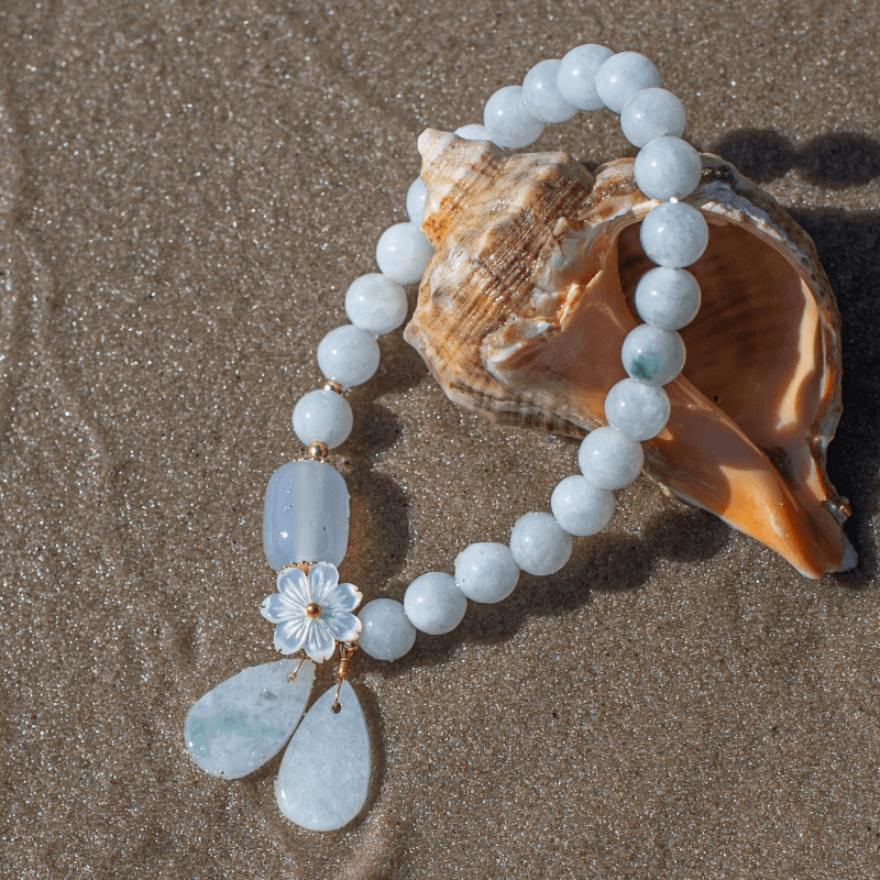Armband aus weißen Perlen mit Muschel und Blumenanhänger auf Sand am Strand.