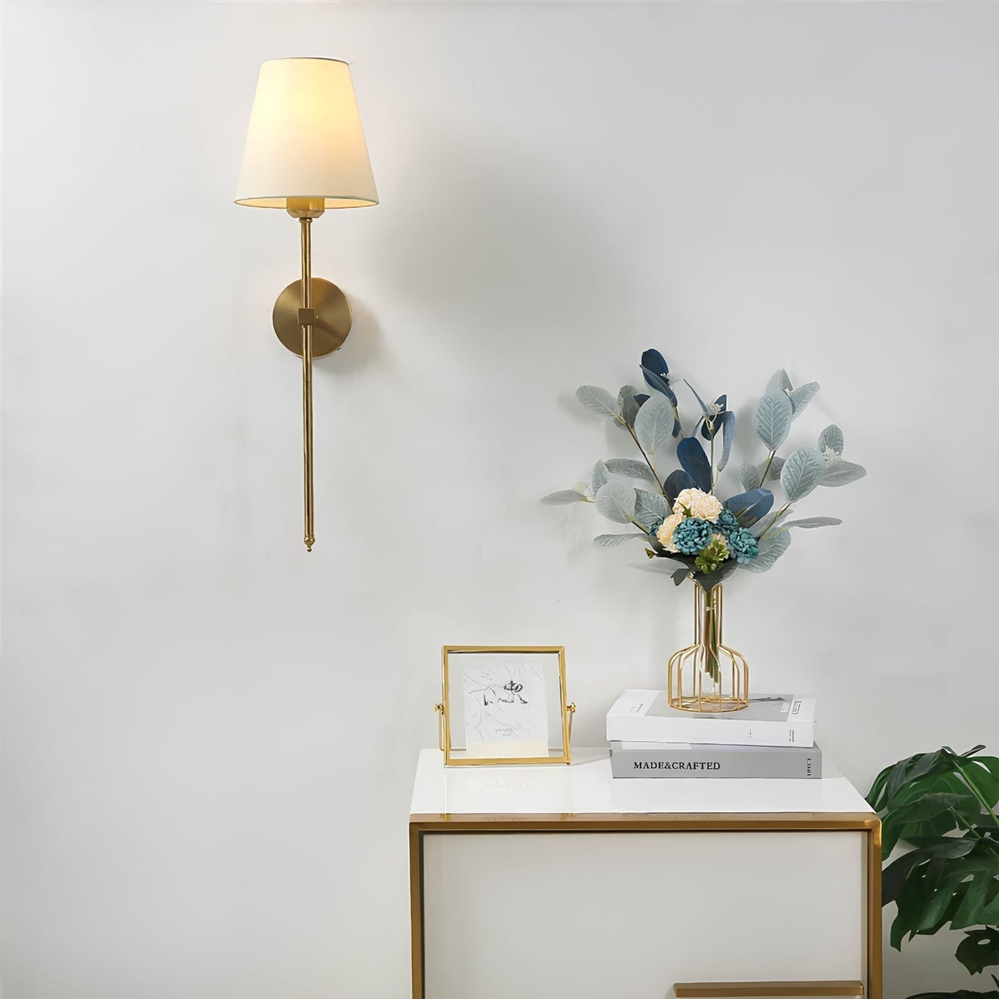 Goldene Wandleuchte mit weißem Lampenschirm neben Tisch mit Blumenstrauß und Dekoration.