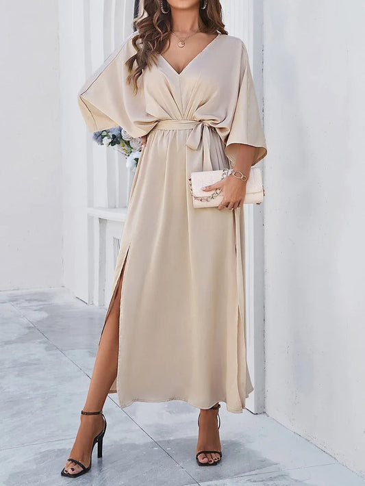 Frau in beigem V-Ausschnitt-Kleid mit Schlitz, hält eine Clutch, trägt High Heels.