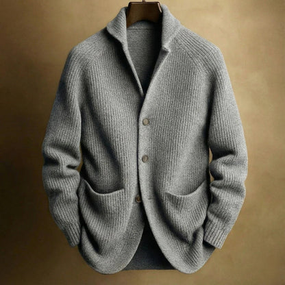 Graue Strickjacke mit grober Rippenstruktur, Raglanärmeln und zwei Taschen, am Bügel vor beigem Hintergrund 5