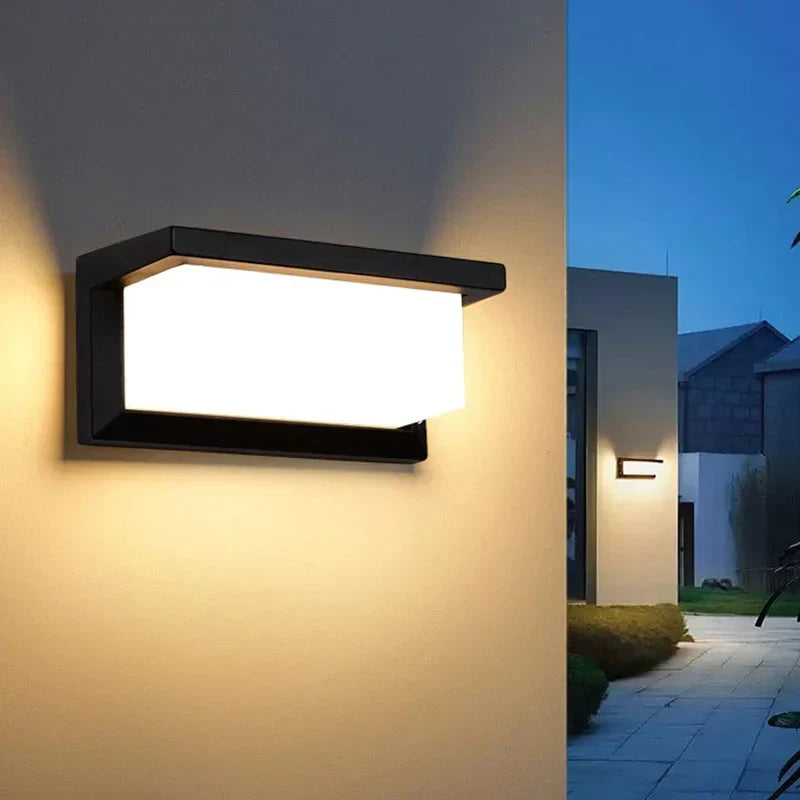 Eingeschaltete Wandlampe an heller Hauswand, im Hintergrund ein Gartenweg in der Abenddämmerung 3