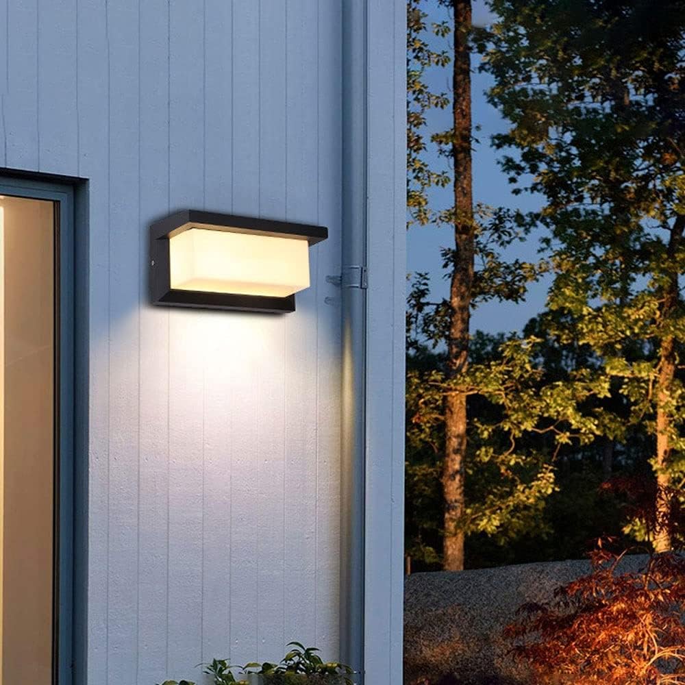Leuchtende Wandlampe an weißer Holzfassade neben Regenrohr, im Hintergrund Bäume bei Dämmerung 6