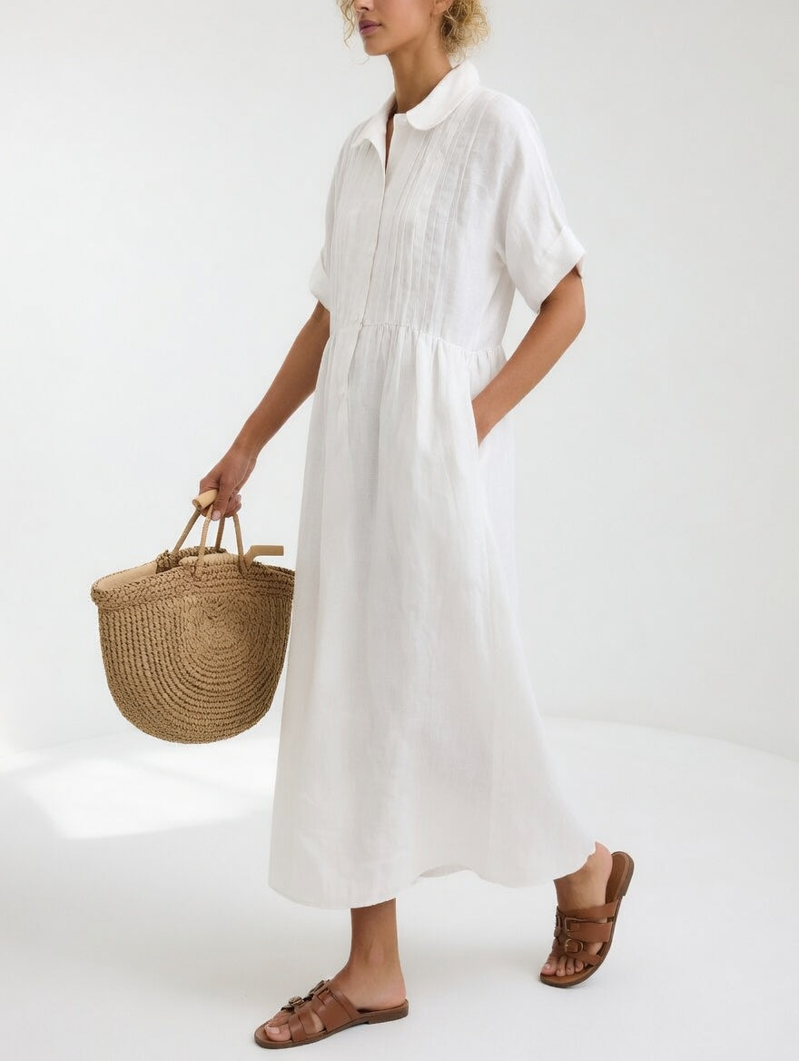 Weißes Maxikleid mit Biesen an der Brust und Bubikragen, seitlich getragen, eine Hand in der Tasche, dazu Korbtasche. 1