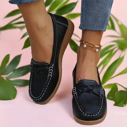 Schwarze Loafer mit geflochtener Zierbrücke und heller Ziernaht, getragen vor rosa Hintergrund mit grünen Blättern und Perlen-Fußkettchen. 2