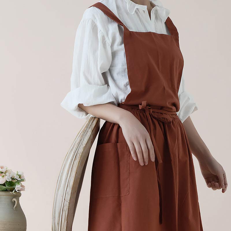 Frau in brauner Schürze und weißem Hemd, steht neben einem Stuhl, Blumen im Hintergrund.