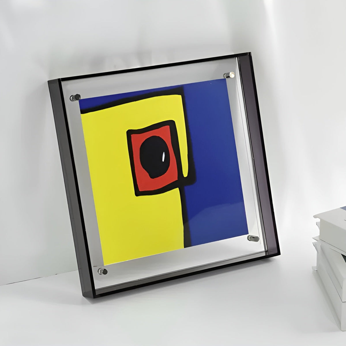 Quadratischer Acrylrahmen mit dunklem Rand lehnt an einer Wand, darin ein abstrakter Druck in Gelb, Blau und Rot; daneben Bücher. 6