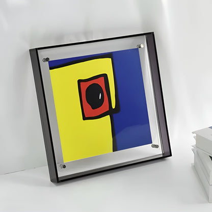 Quadratischer Acrylrahmen mit dunklem Rand lehnt an einer Wand, darin ein abstrakter Druck in Gelb, Blau und Rot; daneben Bücher. 6