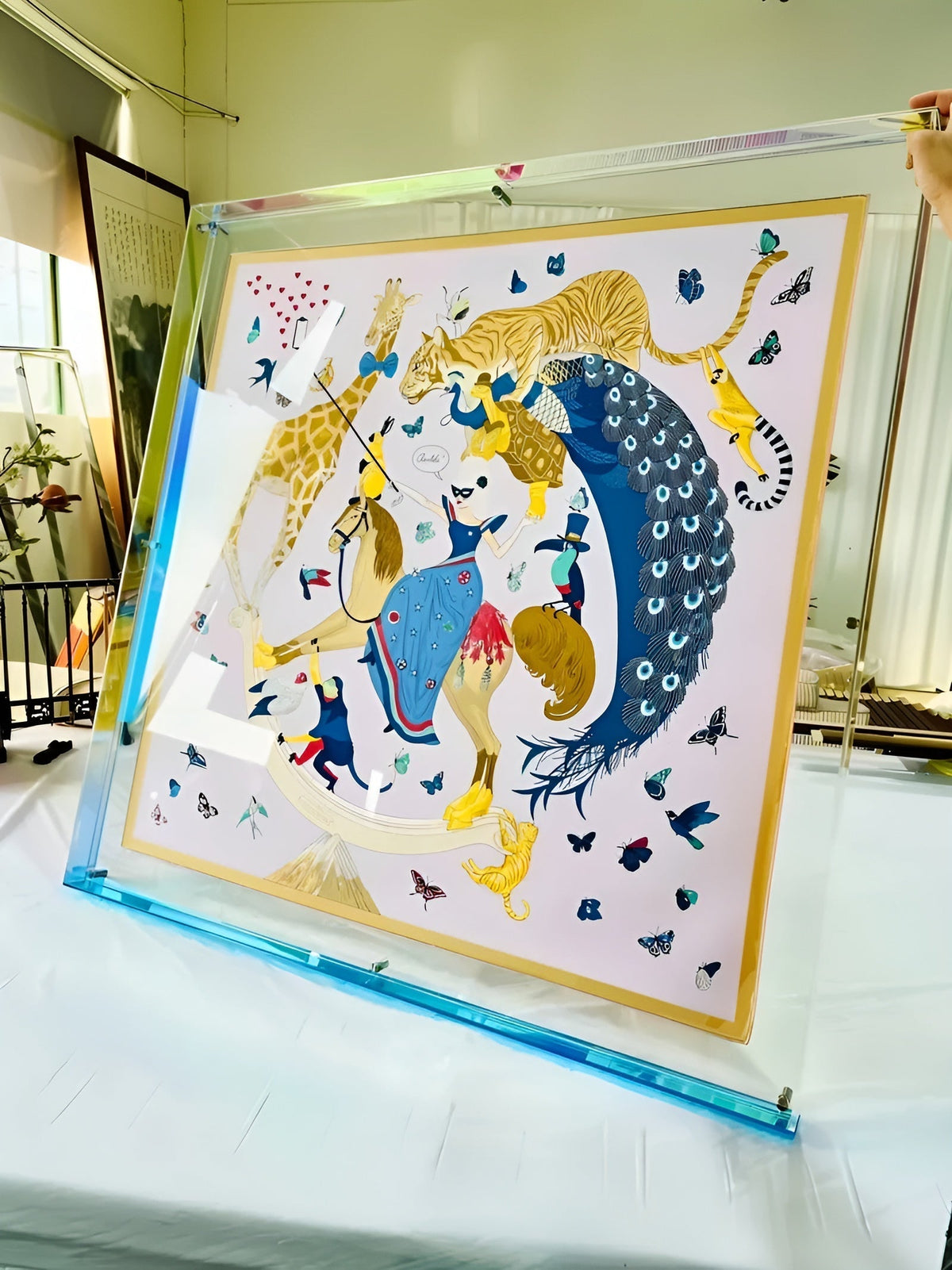 Großer Acrylrahmen mit blau-türkis schimmernder Kante, von einer Hand gehalten, mit gerahmtem Tuch voller Tiere und Schmetterlinge. 17