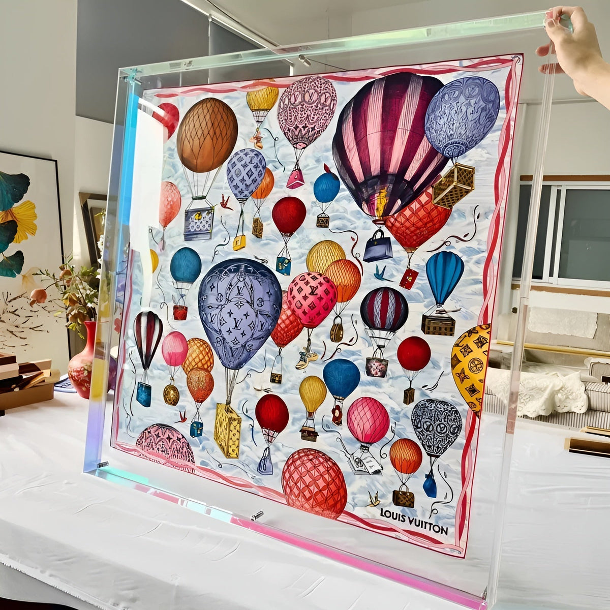 Großer, farblos schimmernder Acrylrahmen, von einer Hand gehalten, mit gerahmtem Seidentuch voller bunter Heißluftballons. 11