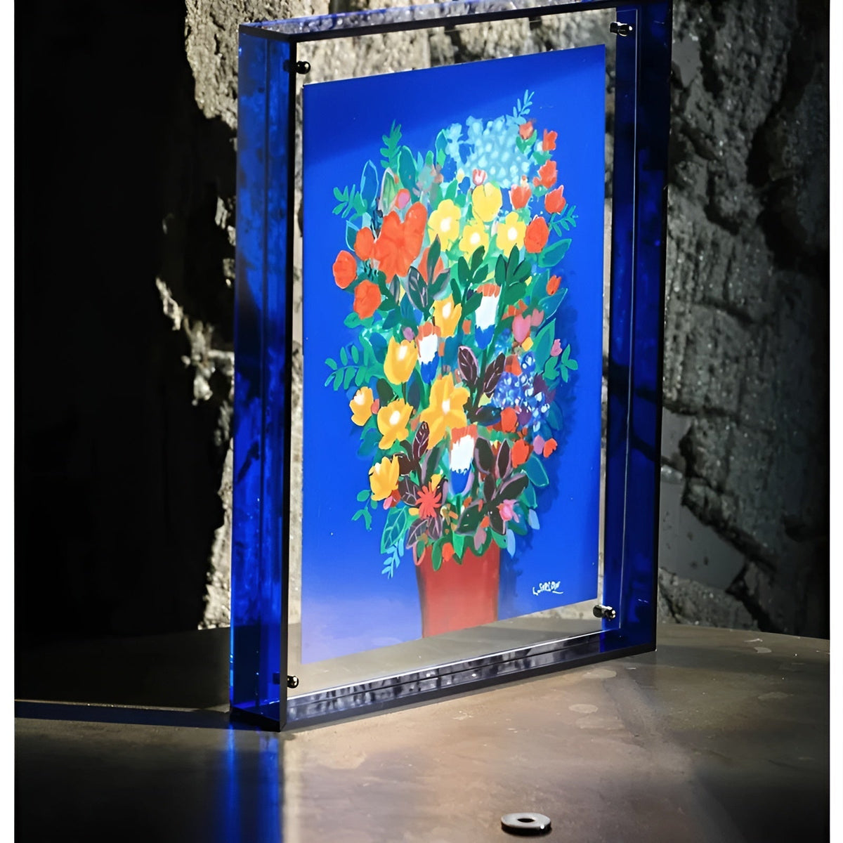 Blauer Acrylrahmen mit Blumenstrauß-Druck steht auf einer Fläche vor einer rauen Steinwand. 4