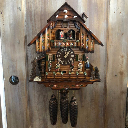 An einer rustikalen Holztür hängende Kuckucksuhr mit bemalten Blumenornamenten, Wasserrad, tanzenden Figuren und drei Tannenzapfengewichten. 12