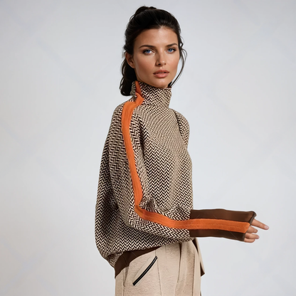 Seitenansicht: Model trägt den Pullover in braun-beigem Fischgrätmuster mit orangefarbenem Streifen über Schulter und Arm, dazu helle Hose. 1
