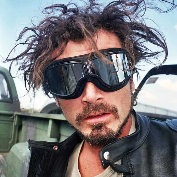 Mann mit zerzaustem Haar und Bart trägt die schwarze Fliegerbrille mit verspiegelten Gläsern, dazu eine Lederjacke. 3