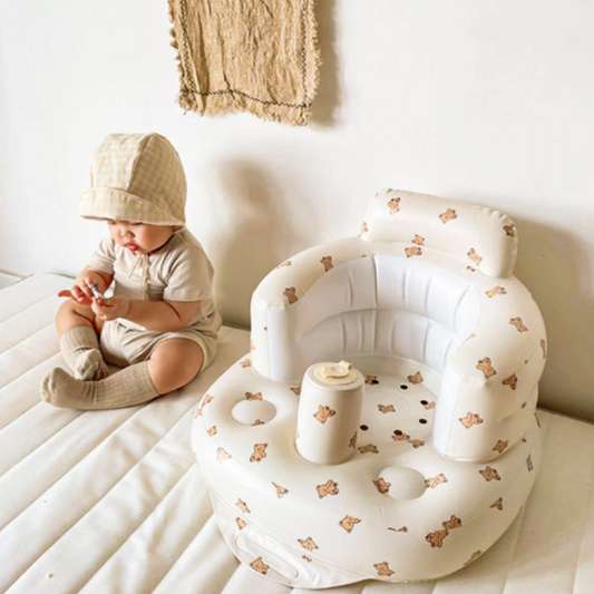 Kleinkind in beigem Outfit neben aufblasbarem Baby-Sitz mit Bärenmuster auf weißer Matratze.