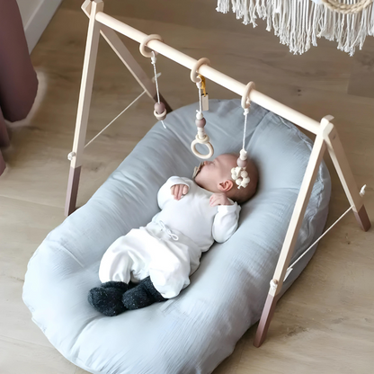 Baby auf grauem Babykissen unter Holzspielbogen mit Hängespielzeug, skandinavisches Design.