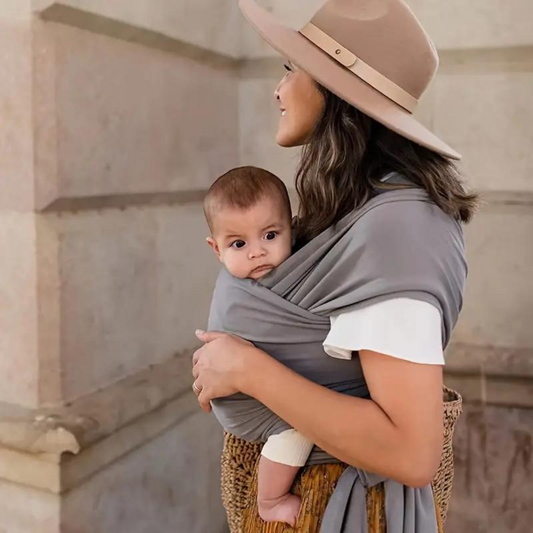 Frau mit Baby in grauem Tragetuch, beige Hut, lächelnd, draußen, Mutter-Kind-Bindung.