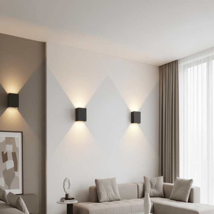 Drei schwarze, würfelförmige Wandleuchten strahlen warmes Licht nach oben und unten auf eine helle Wand über einem beigen Sofa. 1