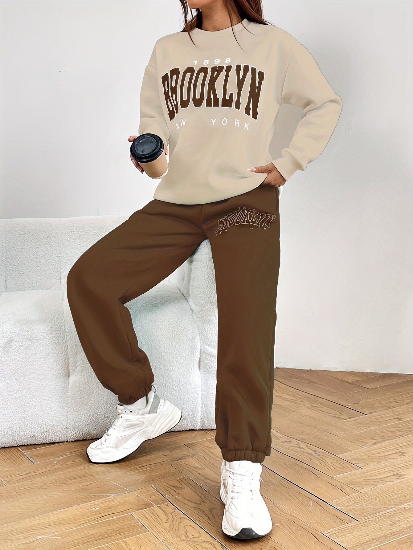 Frau in beigem Brooklyn-Sweatshirt und brauner Jogginghose, hält schwarzen Becher, weiße Sneaker.